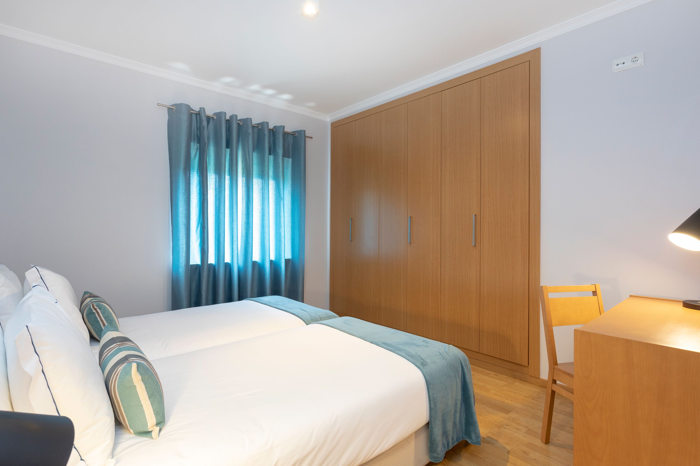 A hotel room with a bed, a wooden wardrobe, a desk with a chair, and a window with curtains.