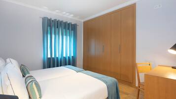 A hotel room with a bed, a wooden wardrobe, a desk with a chair, and a window with curtains.