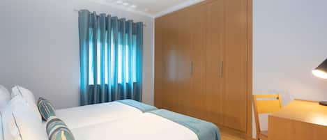A hotel room with a bed, a wooden wardrobe, a desk with a chair, and a window with curtains.