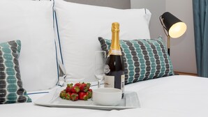 Una cama bien hecha con almohadillas blancas y azul verdosas, una botella de champán, una copa y una bandeja con fresas y un cuenco.