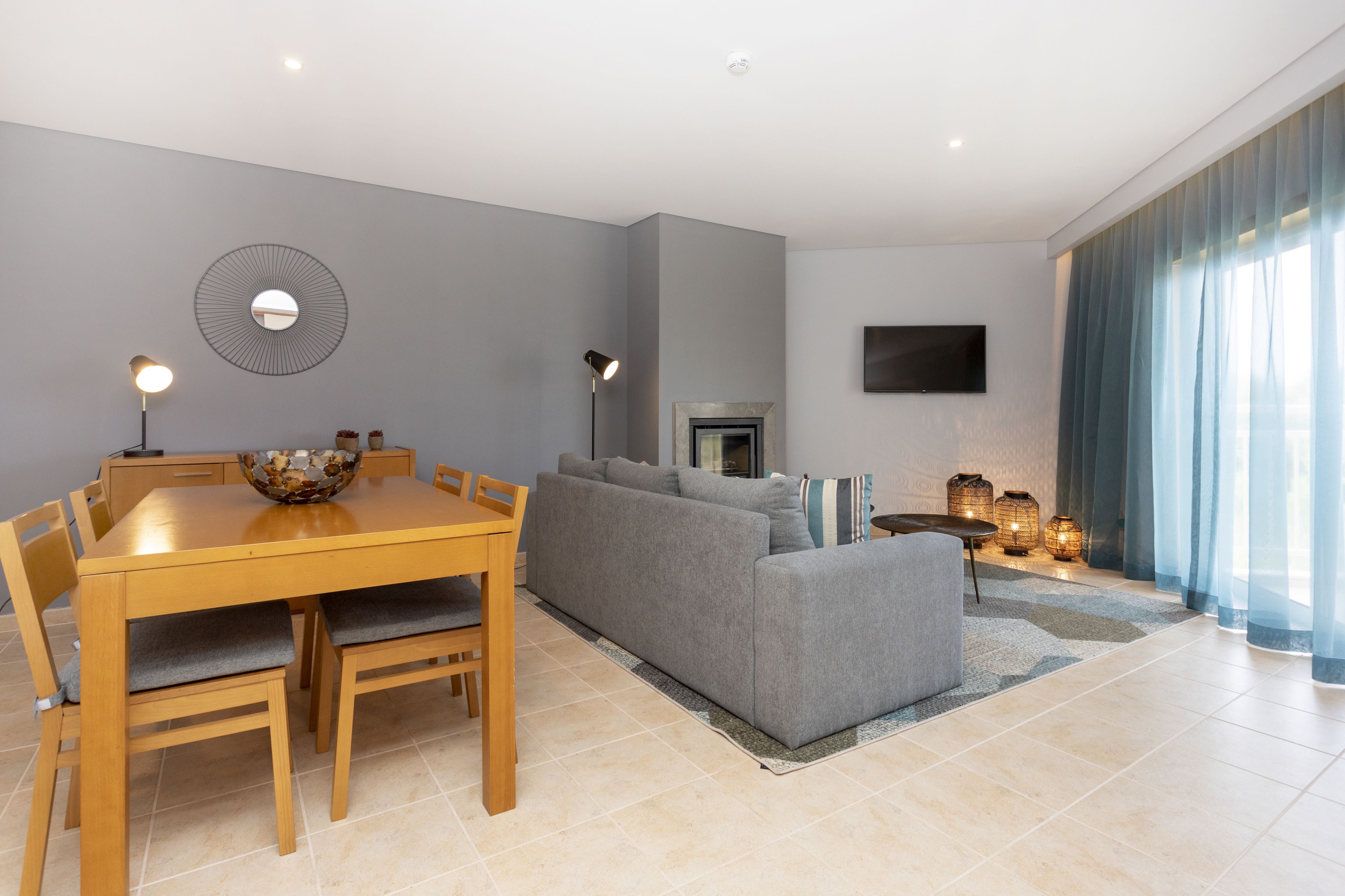 A modern living room with a grey sofa, wooden dining table, and chairs. There is a flat-screen TV mounted on the wall and a fireplace.