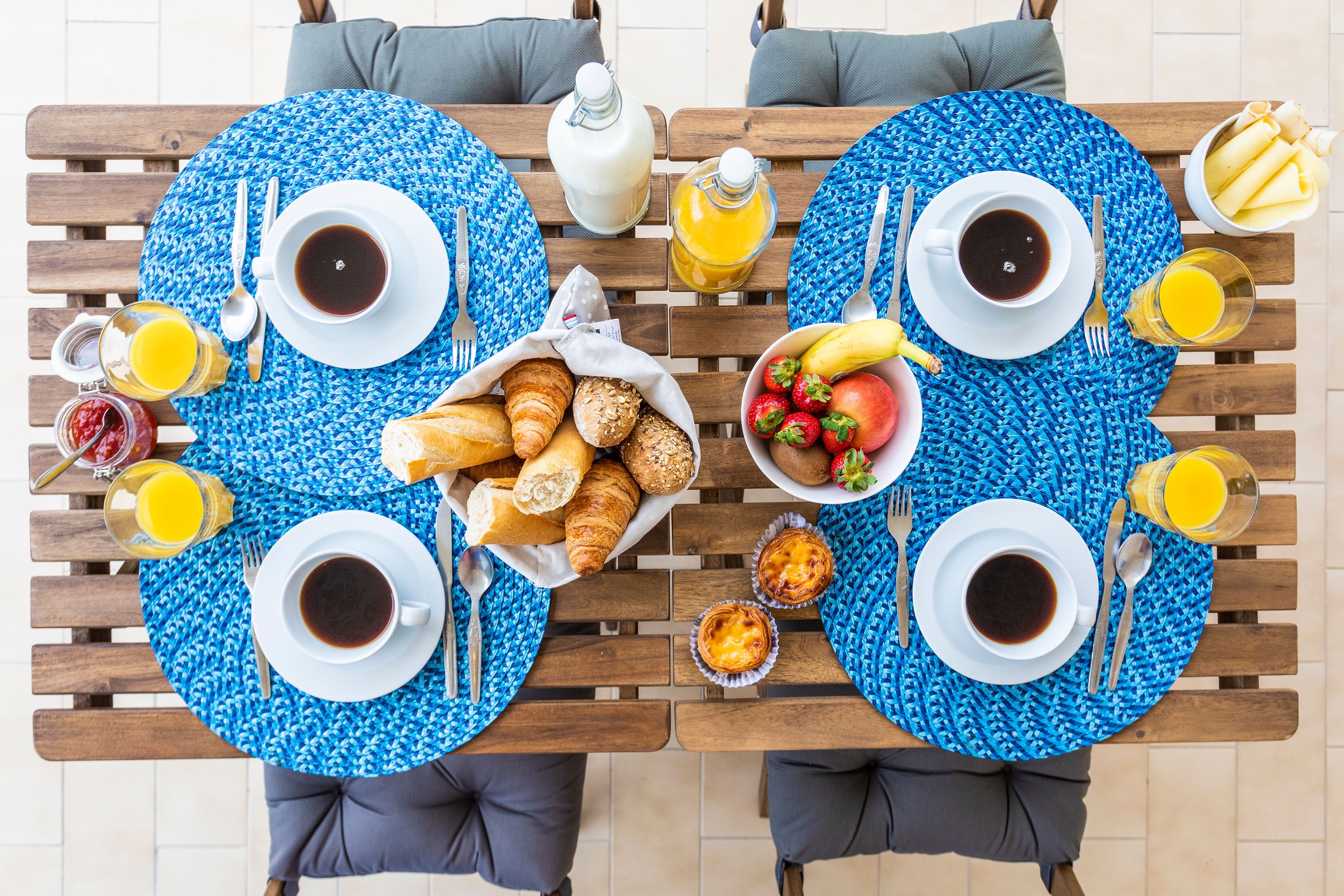 A dining table with blue placemats, two sets of cutlery, plates, cups, and a variety of breakfast items including pastries, fruits, and beverages.