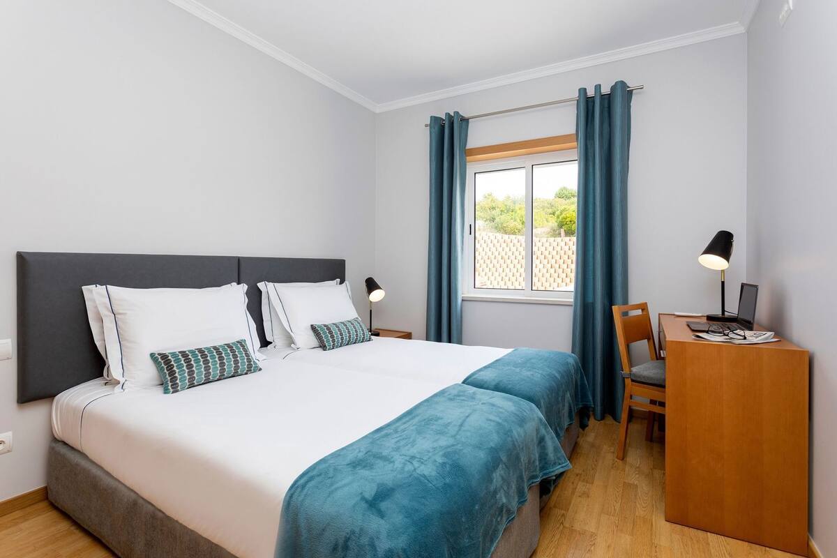 A hotel room with a bed, a desk, a chair, a lamp, and a window with curtains.