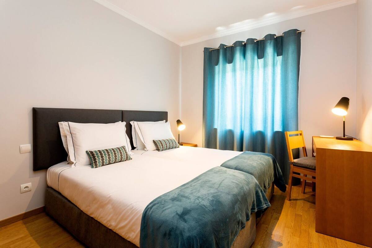 A hotel room with a bed, a desk, a chair, a lamp, and a window with curtains.