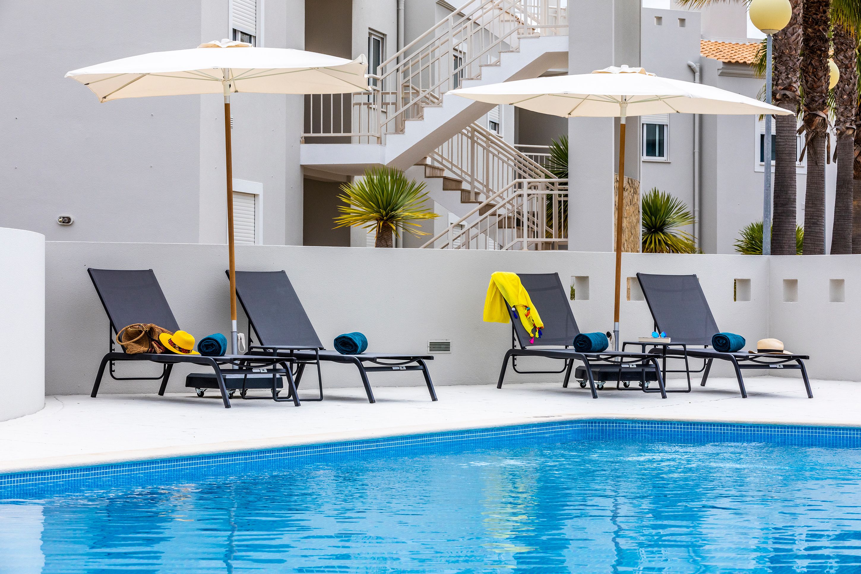 A poolside area with lounge chairs and umbrellas, a tiled pool, and a building with balconies and stairs in the background.
