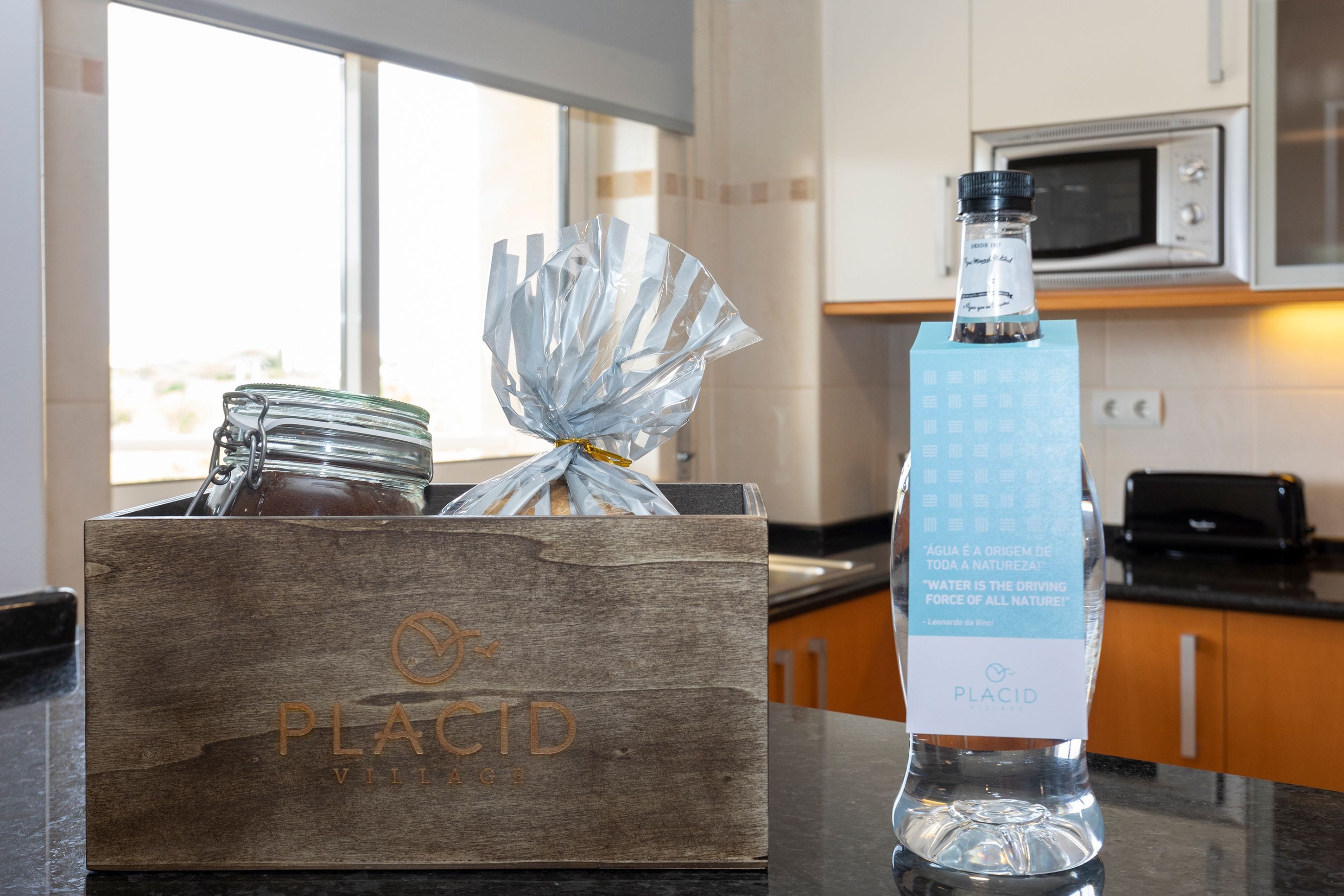 Un comptoir de cuisine sur lequel se trouve une boîte en bois étiquetée « PLACID VILLAGE », contenant un bocal en verre et un objet emballé. On peut également apercevoir une bouteille d’eau « PLACID ».