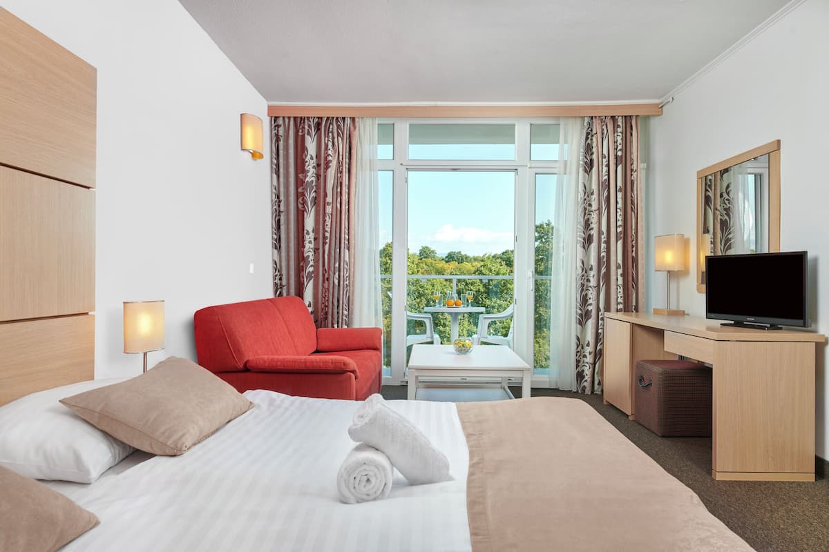 Ein Hotelzimmer mit einem Bett, einem roten Sessel, einem Schreibtisch mit Fernseher und einem Balkon mit Tisch und Stühlen.