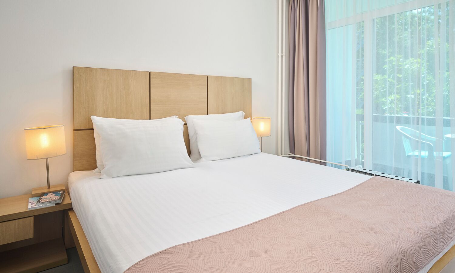 Et hotelværelse med en stor seng, to sengeborde, lamper og udsigt til det fri gennem store vinduer.