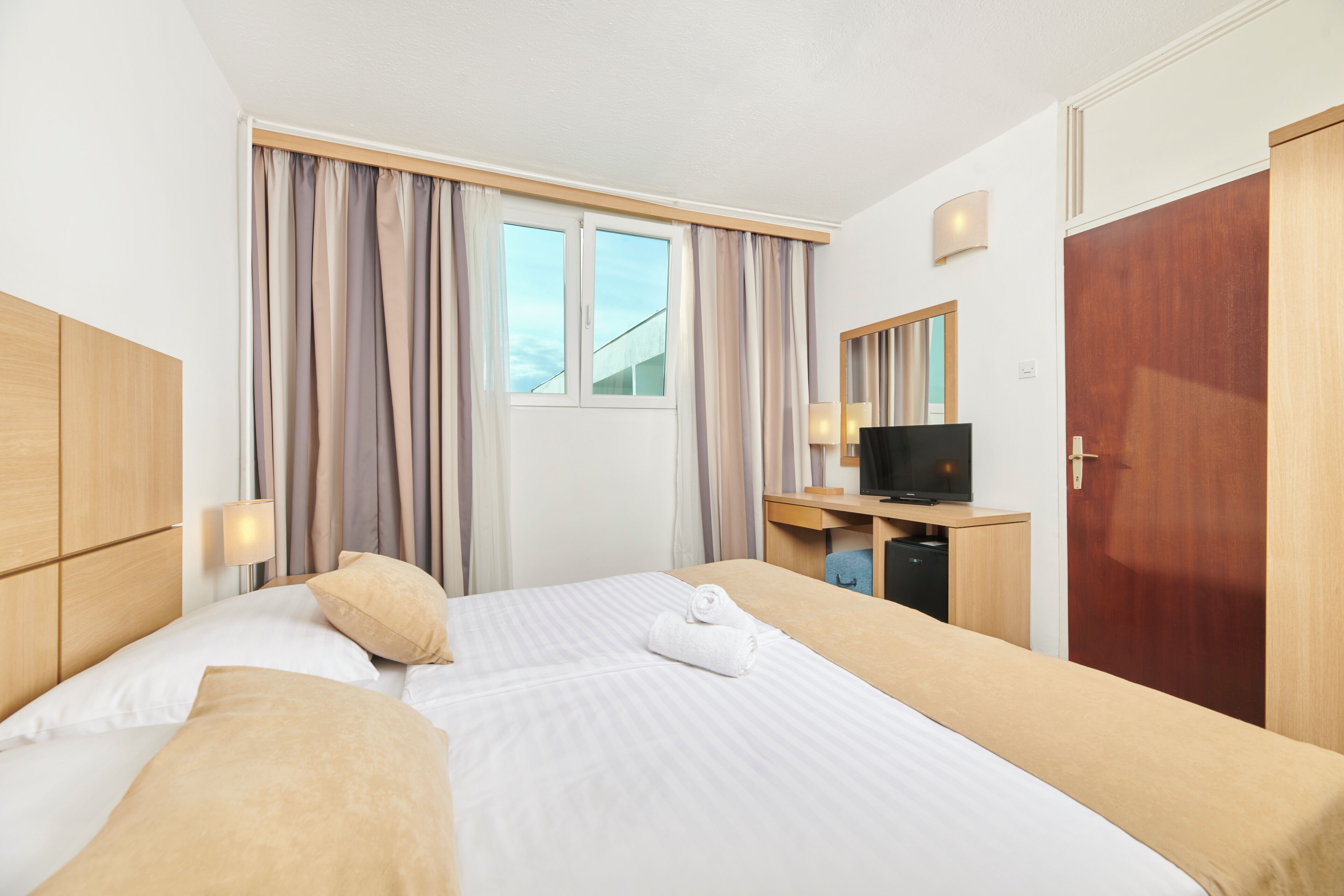 Une chambre d’hôtel avec un grand lit, un bureau avec une télévision, une armoire et une fenêtre avec des rideaux.