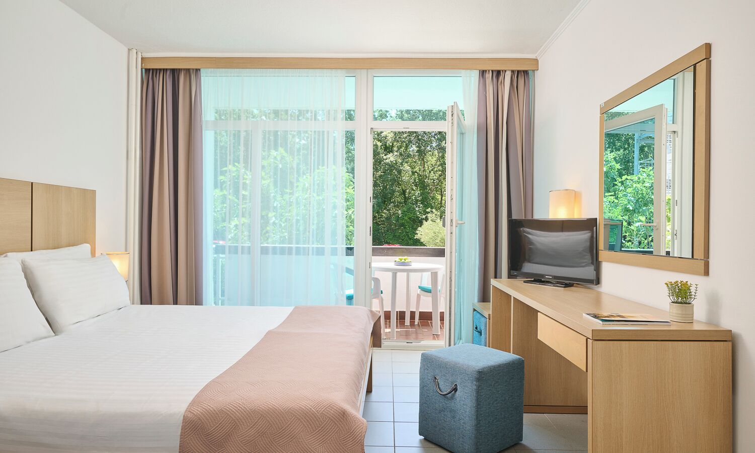 Une chambre d’hôtel comprenant un lit, un bureau avec une télévision, une petite table avec des chaises et un balcon donnant sur la forêt.