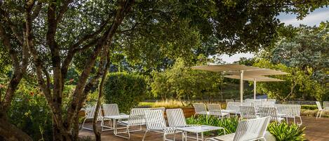 An outdoor seating area with white metal chairs and a table, surrounded by greenery and trees.