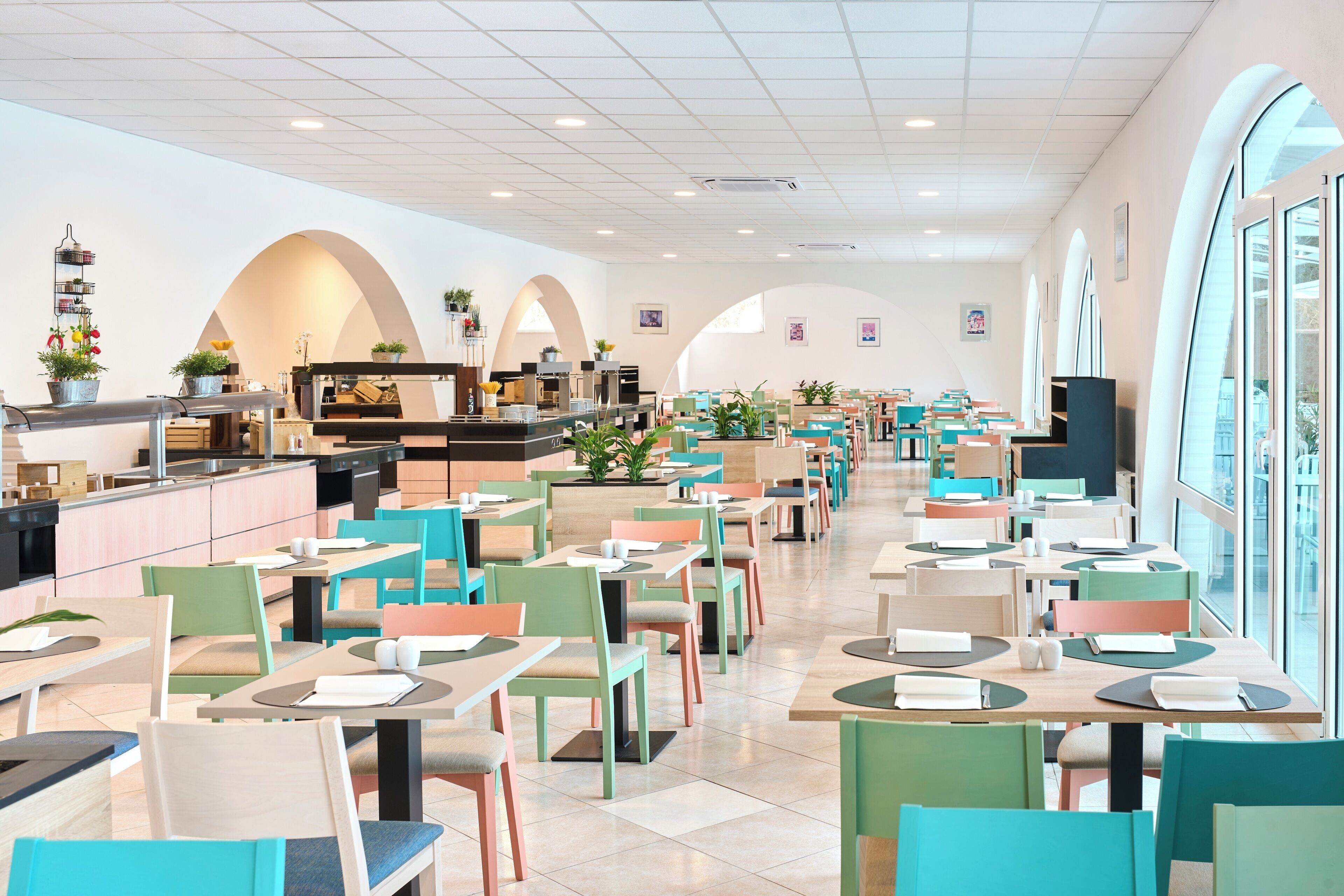 Un restaurante amplio con múltiples mesas y sillas, una barra con máquina de café y ventanas grandes que dejan entrar luz natural.