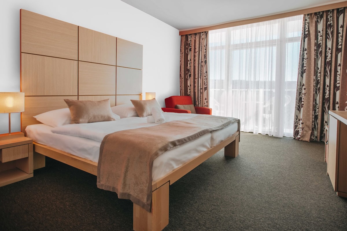 Ein Hotelzimmer mit einem großen Bett, einem Fernseher auf einer Holzkabine, einem roten Sessel und einem großen Fenster mit Vorhängen.