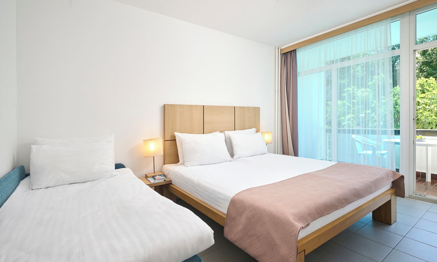 Ein Hotelzimmer mit zwei Betten, einem Holzkopfende, einem großen Fenster mit Vorhängen und einem Balkon mit Sitzmöbeln.