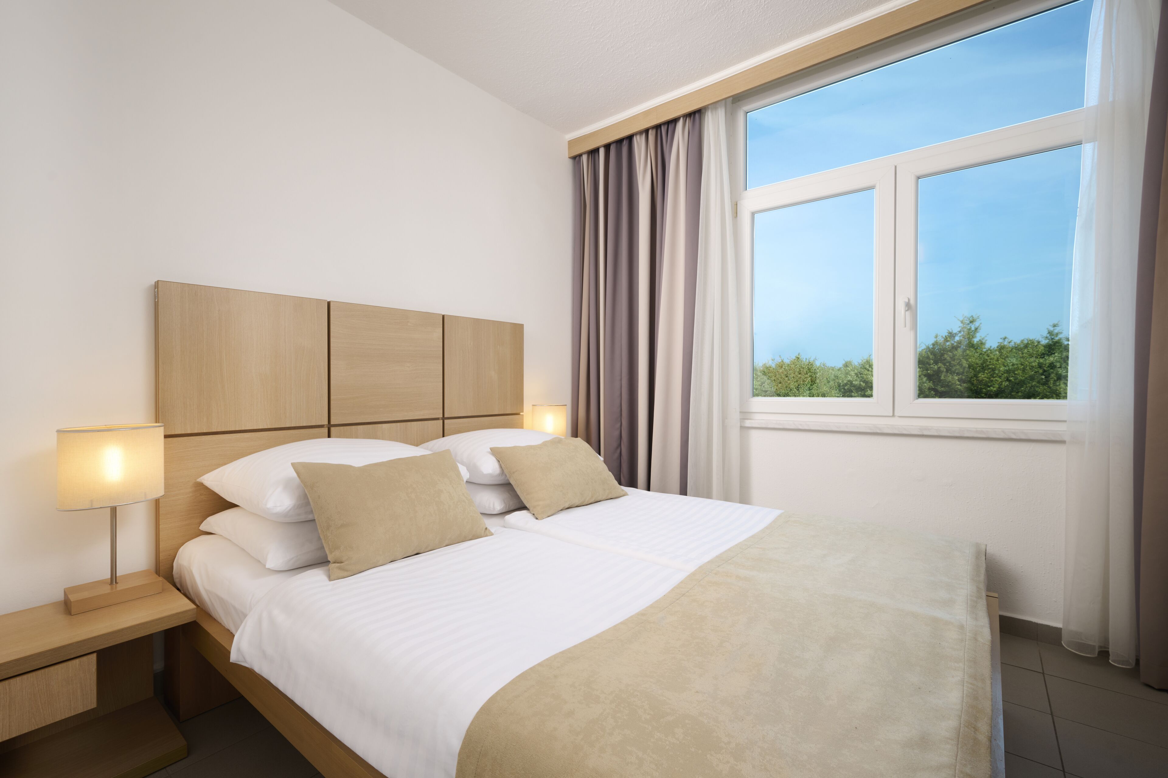 Cama bem arrumada com cabeceira de madeira, duas luminárias de apoio e vista para o verde através de uma janela com cortinas.