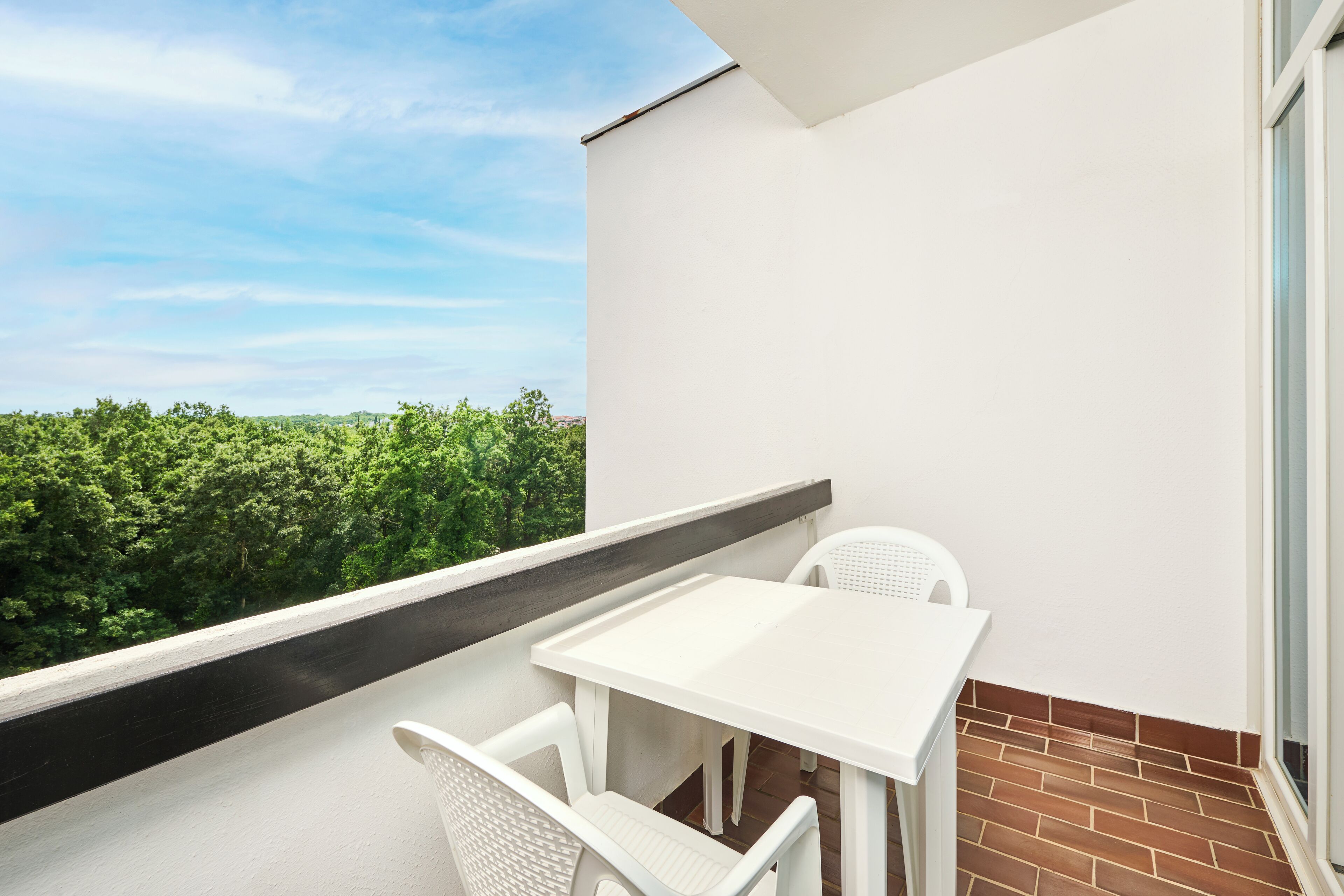 A balcony with a white table and chairs, a brick floor, and a view of green trees.