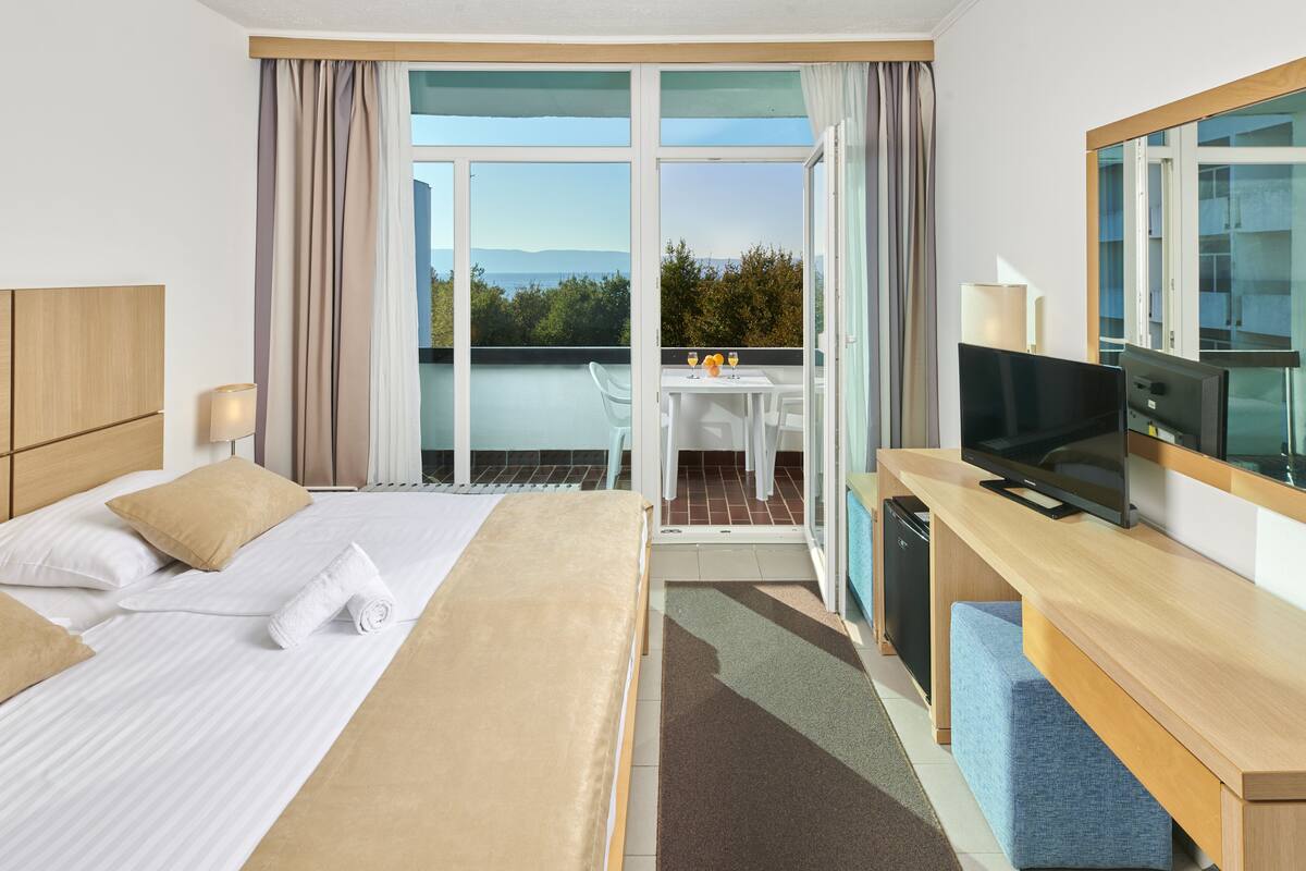 Ein Hotelzimmer mit Bett, Schreibtisch mit Fernseher, kleinem Tisch mit Gläsern und Blick ins Freie.