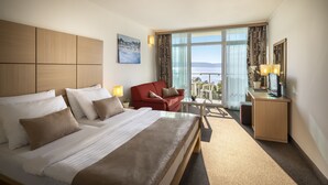 A hotel room with a large bed, a red armchair, a small table, and a desk with a lamp. There is a window with a view of the sea. - Magal Maradiso Hotel by Aminess  (Omisalj)