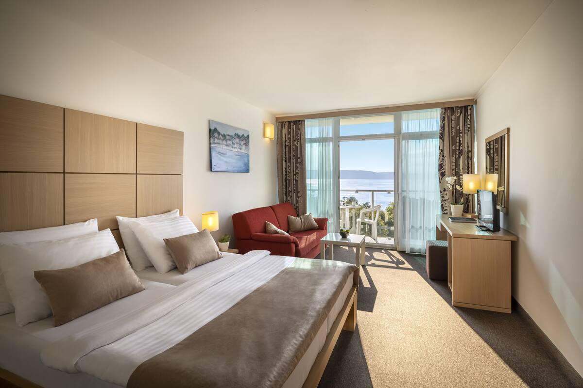 Ein Hotelzimmer mit einem großen Bett, einem roten Sessel, einem kleinen Tisch und einem Schreibtisch mit Lampe. Ein Fenster bietet Blick auf das Meer.