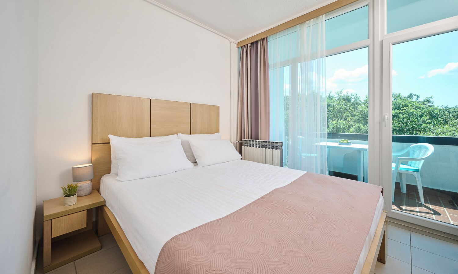 Habitación de hotel con cama, mesita de noche, lámpara, ventana con cortinas y vista a la vegetación.