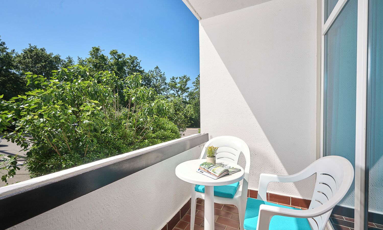 A balcony with a white chair, a small table with a book, and a view of greenery.