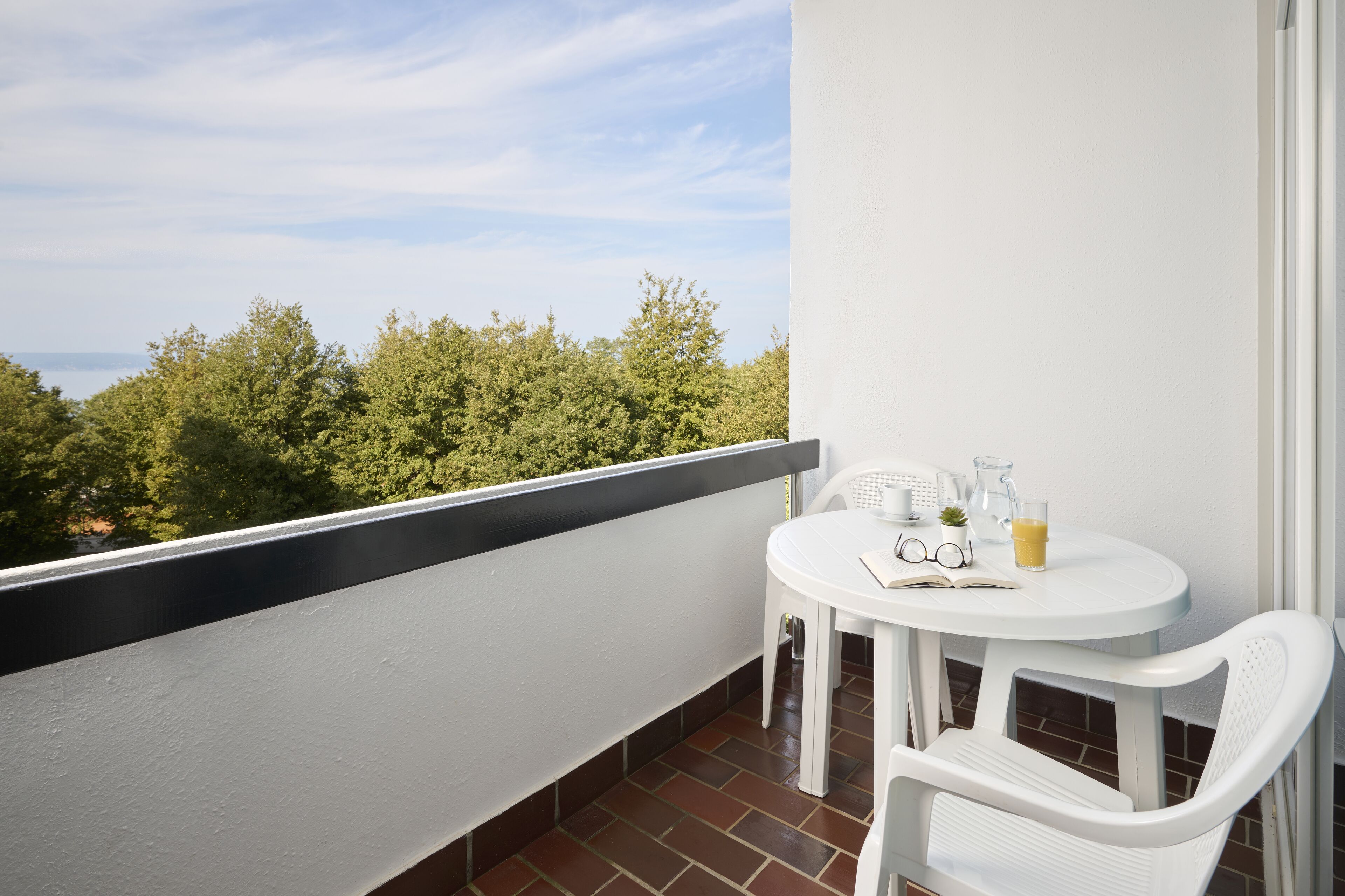 Een balkon met een witte tafel, een stoel, een boek, een glas sinaasappelsap en een waterkan.