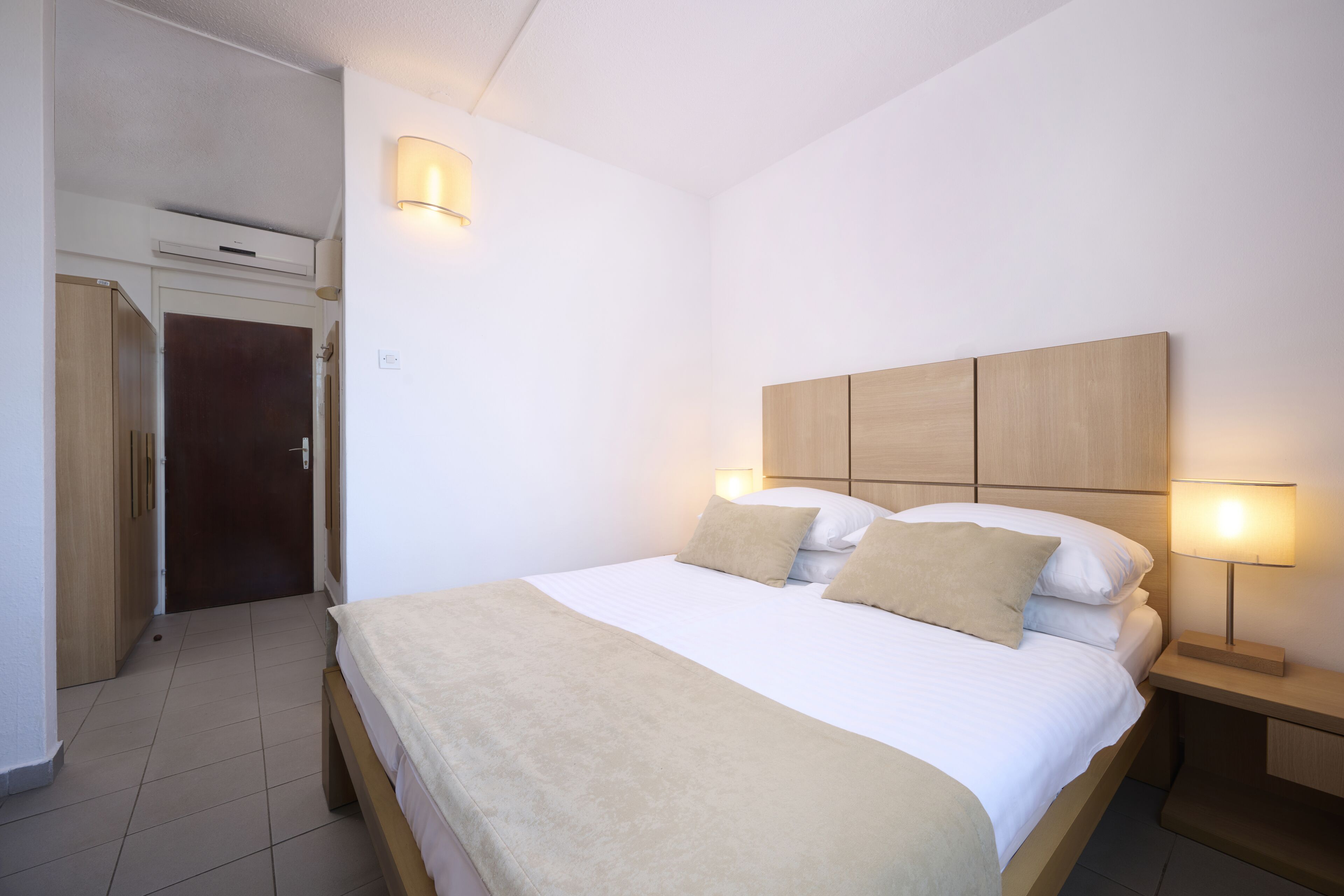 Un dormitorio con cama, lámparas de noche, cabecero de madera y una puerta que da a otra habitación.
