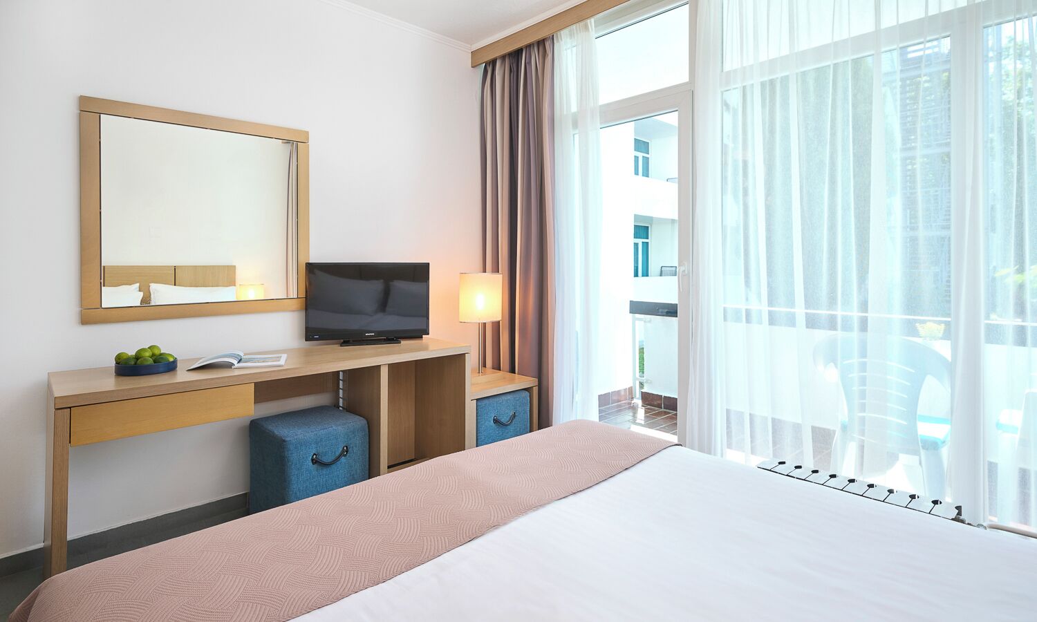 Habitación de hotel con cama, televisor sobre un escritorio de madera, lámpara y vistas al exterior.