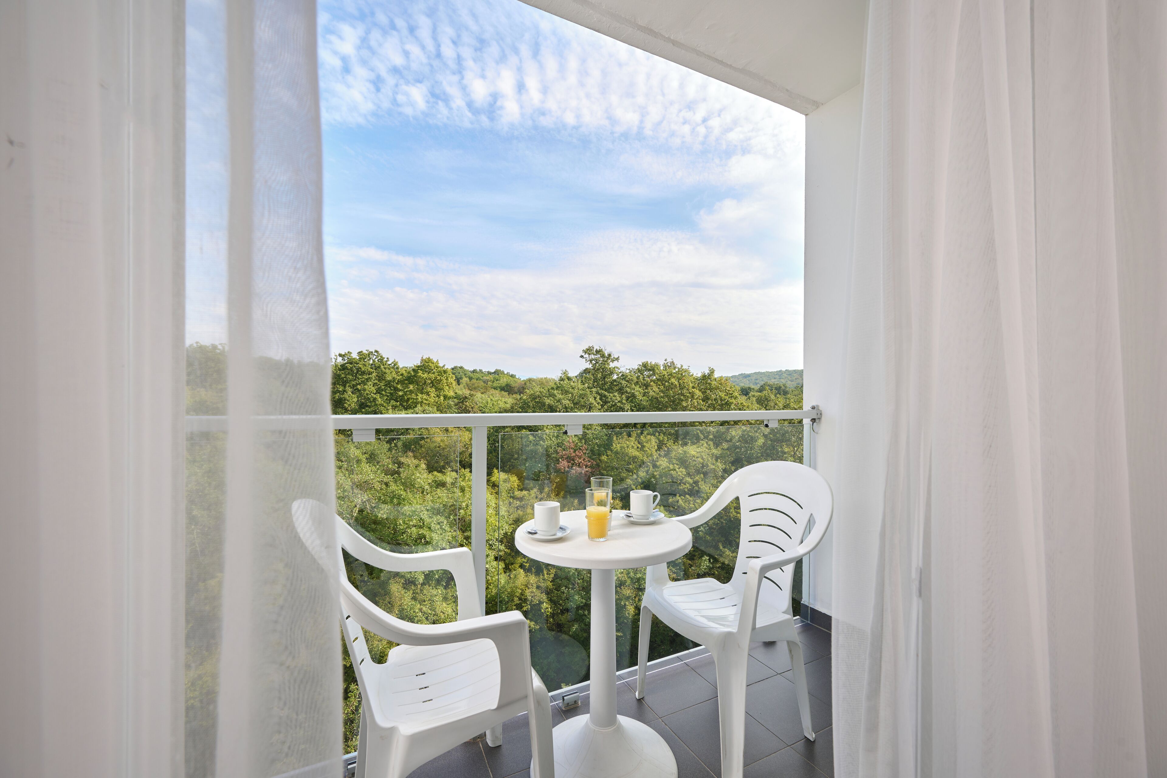 Ein Balkon mit zwei weißen Plastikstühlen und einem kleinen runden Tisch, auf dem ein Glas Orangensaft und zwei Tassen stehen.