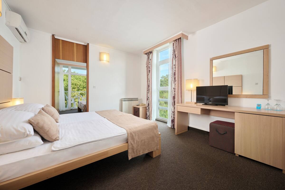 Ein Hotelzimmer mit Bett, Schreibtisch, Fernseher, Spiegel und einem fensterbepflanzten Fenster.