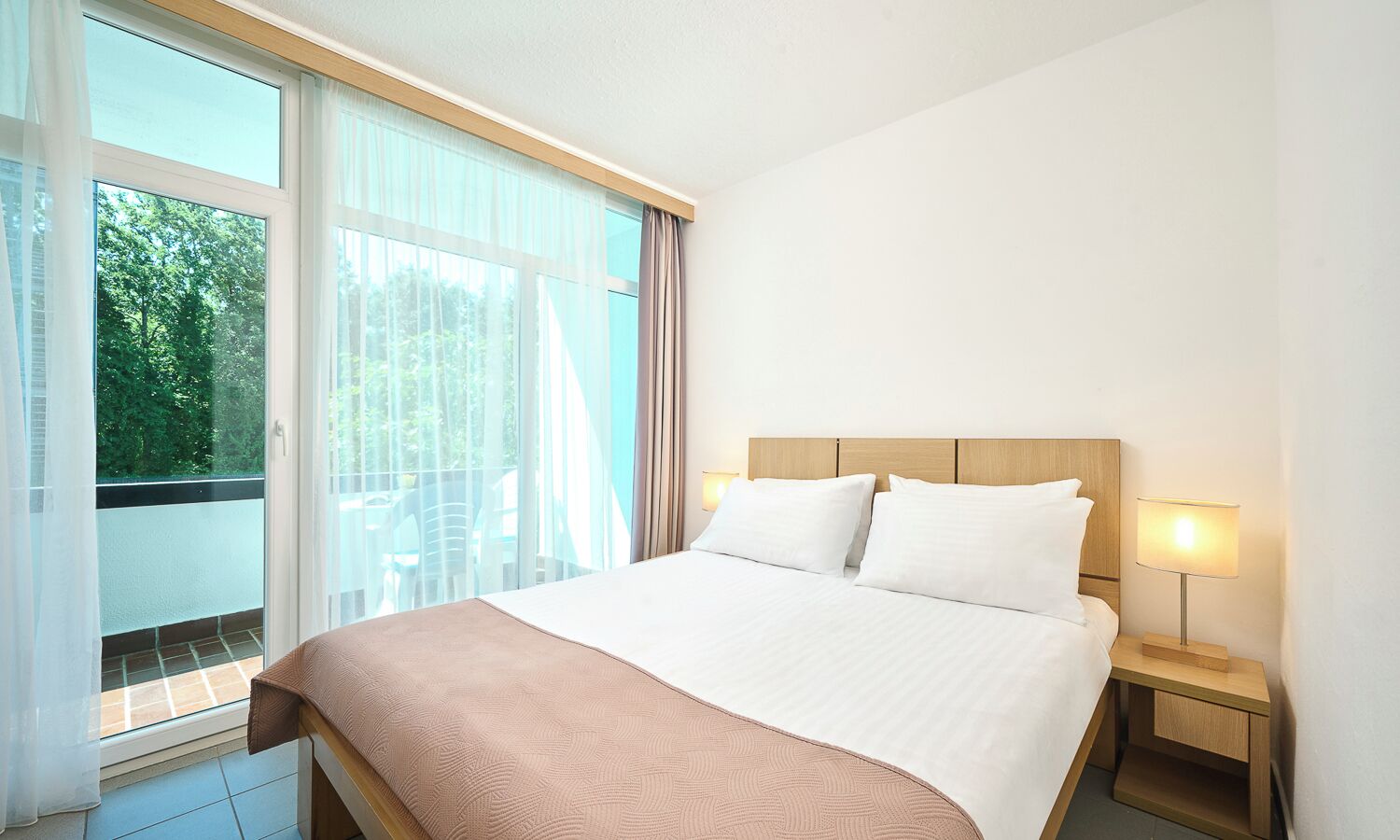 A hotel room with a bed, a nightstand with a lamp, and a view of greenery through the window.