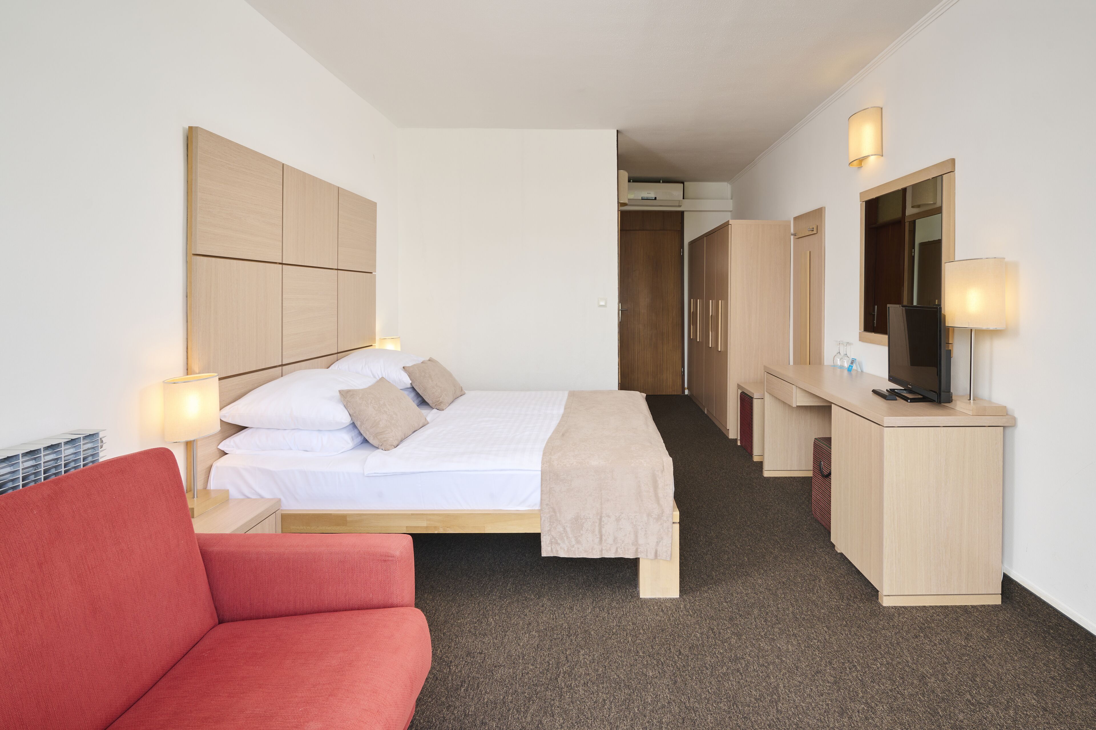 A hotel room with a bed, a desk with a computer, a TV, and a red sofa.