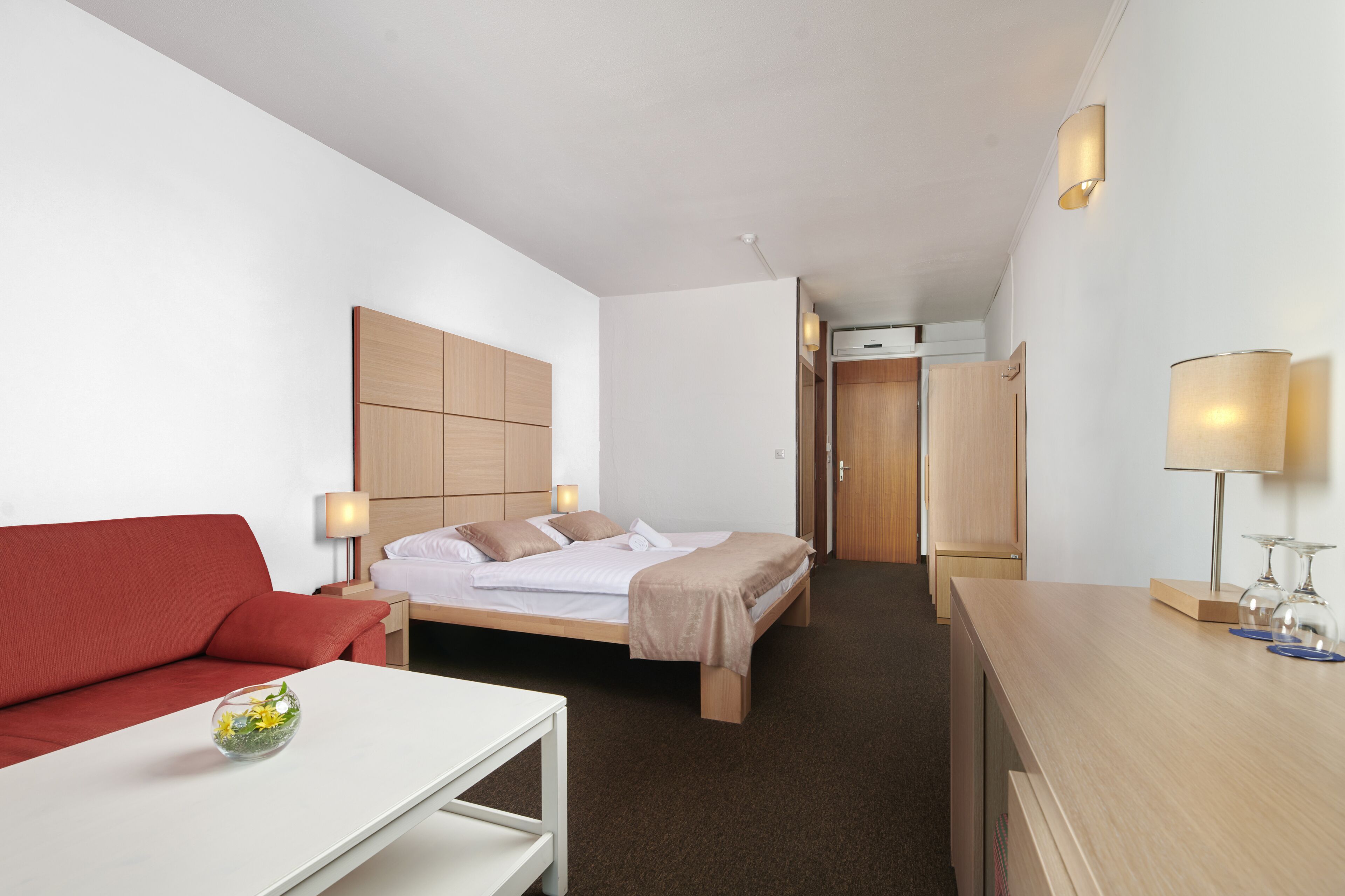 Ett hotellrum med en säng, en röd soffa, ett vitt bord och ett skrivbord med en lampa.