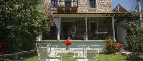Una casa de piedra con balcón, un porche cubierto y un jardín con sillas y flores.