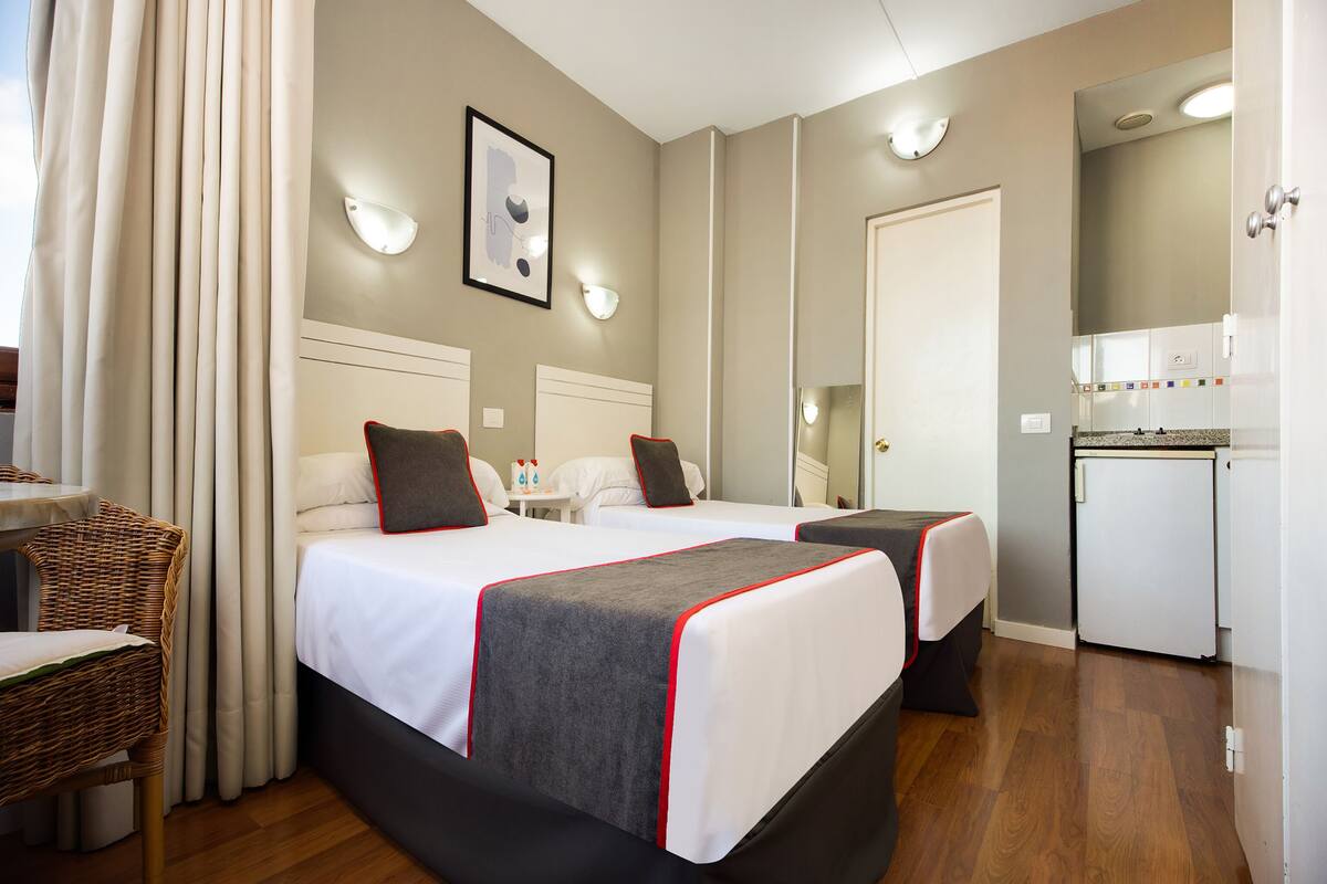 A hotel room with two beds, a small table, a chair, a mirror, and a kitchenette.