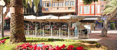 An outdoor café with tables and umbrellas, surrounded by greenery and buildings in the background.