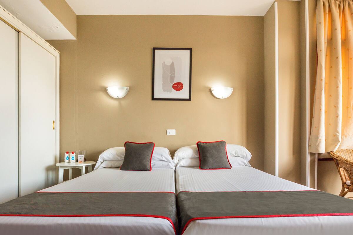 A hotel room with two beds, a framed picture on the wall, and two wall-mounted lamps.