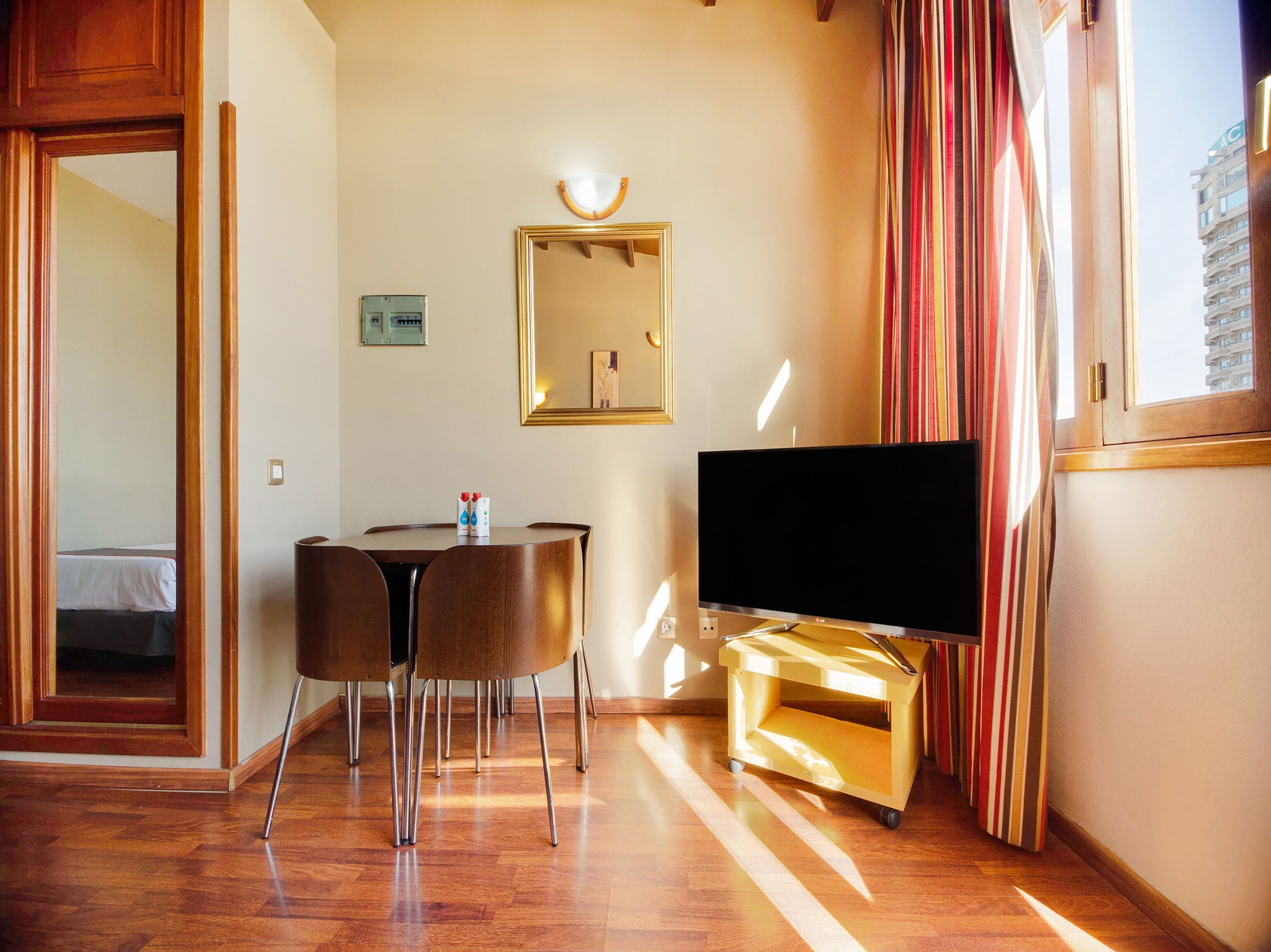 A hotel room with a wooden floor, a TV on a stand, a dining table with chairs, a mirror on the wall, and a window with curtains.