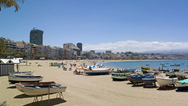 Een strand met veel boten en parasols, een heldere lucht en gebouwen op de achtergrond.