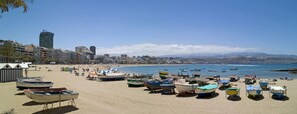 Ein Strand mit zahlreichen Booten und Sonnenschirmen, einem wolkenlosen Himmel und Gebäuden im Hintergrund.
