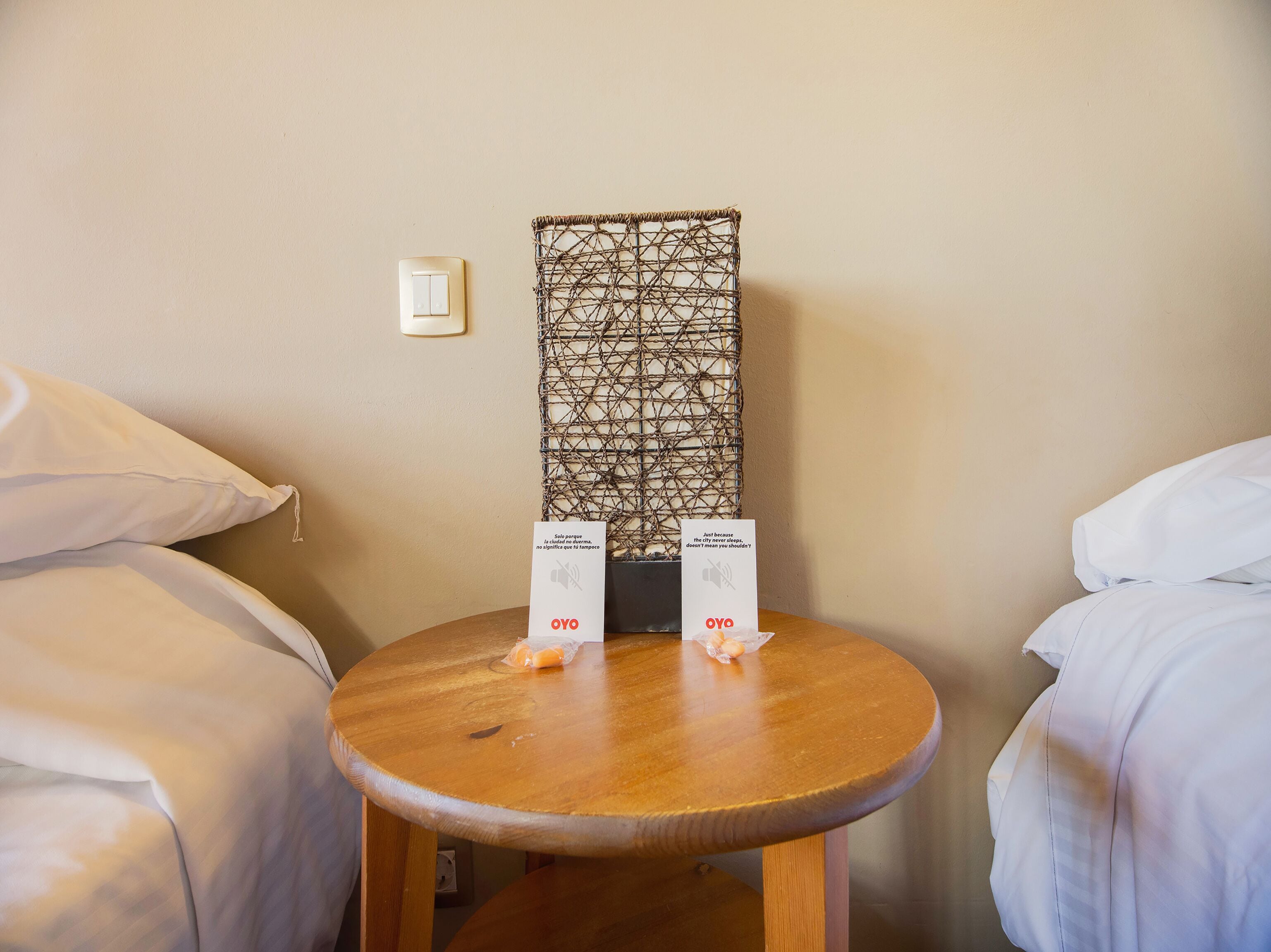 A hotel room with a wooden side table, two white pillows, a woven wall decoration, and two signs with the text "OYO" and "Just because the city never sleeps, doesn't mean you shouldn't?"