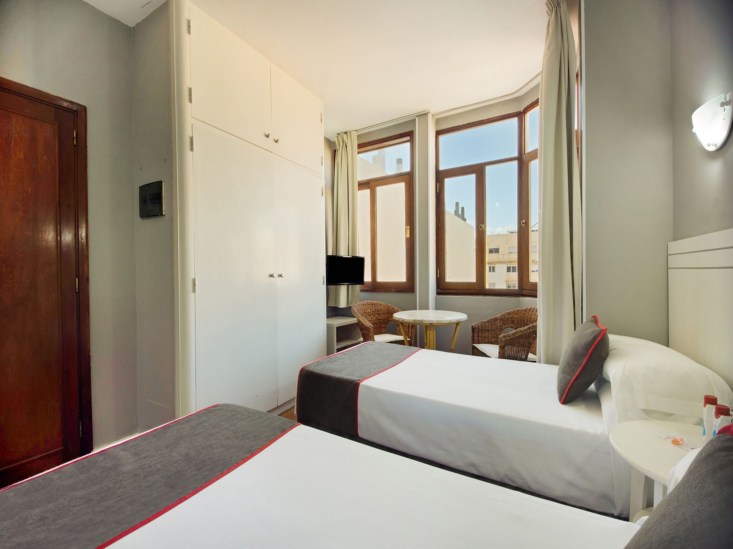 A hotel room with two beds, a television, a small table, wicker chairs, and a view of buildings through large windows.