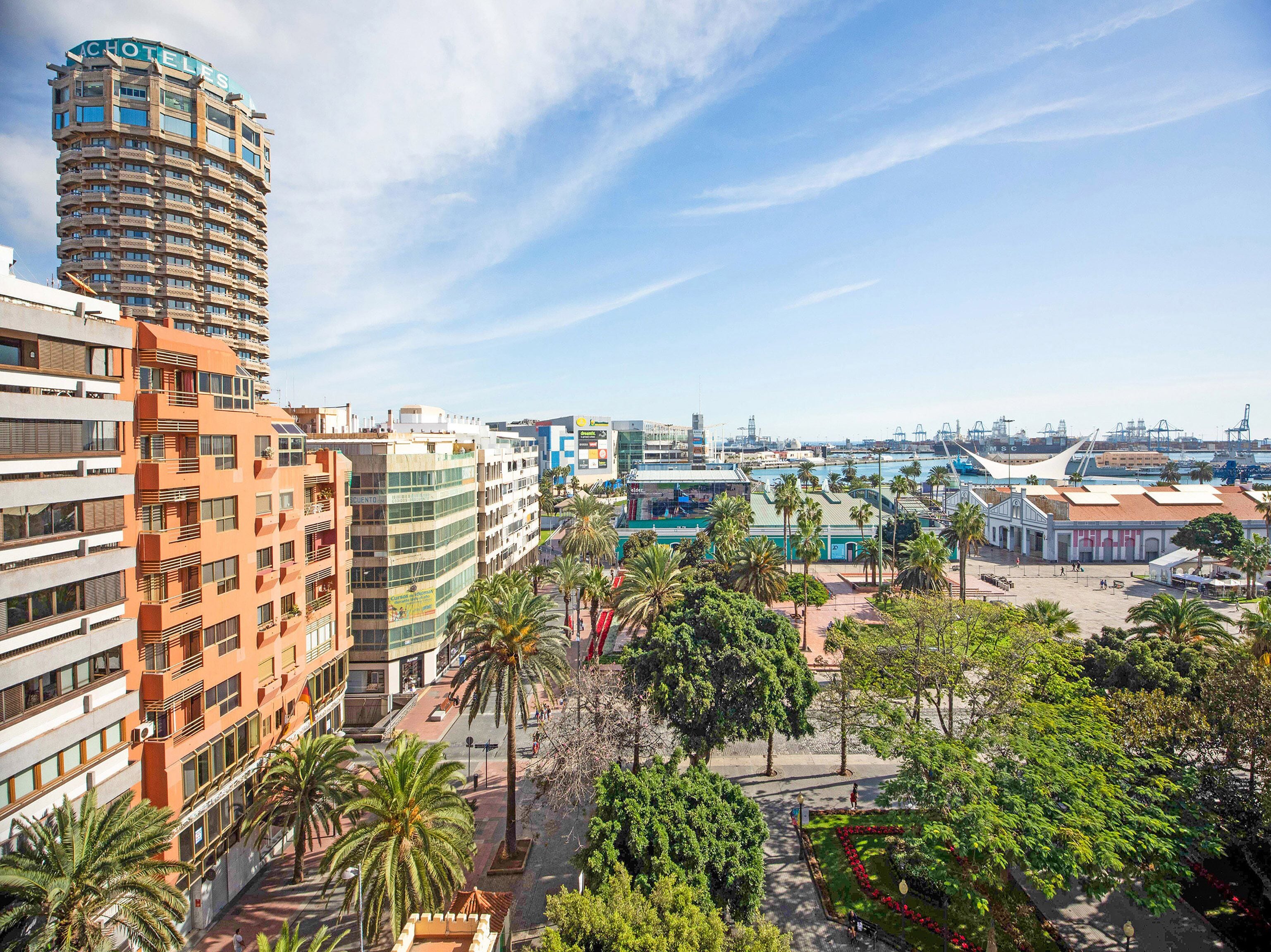 A high-rise hotel with multiple balconies, a central courtyard with palm trees, and a view of the marina and port in the background.
