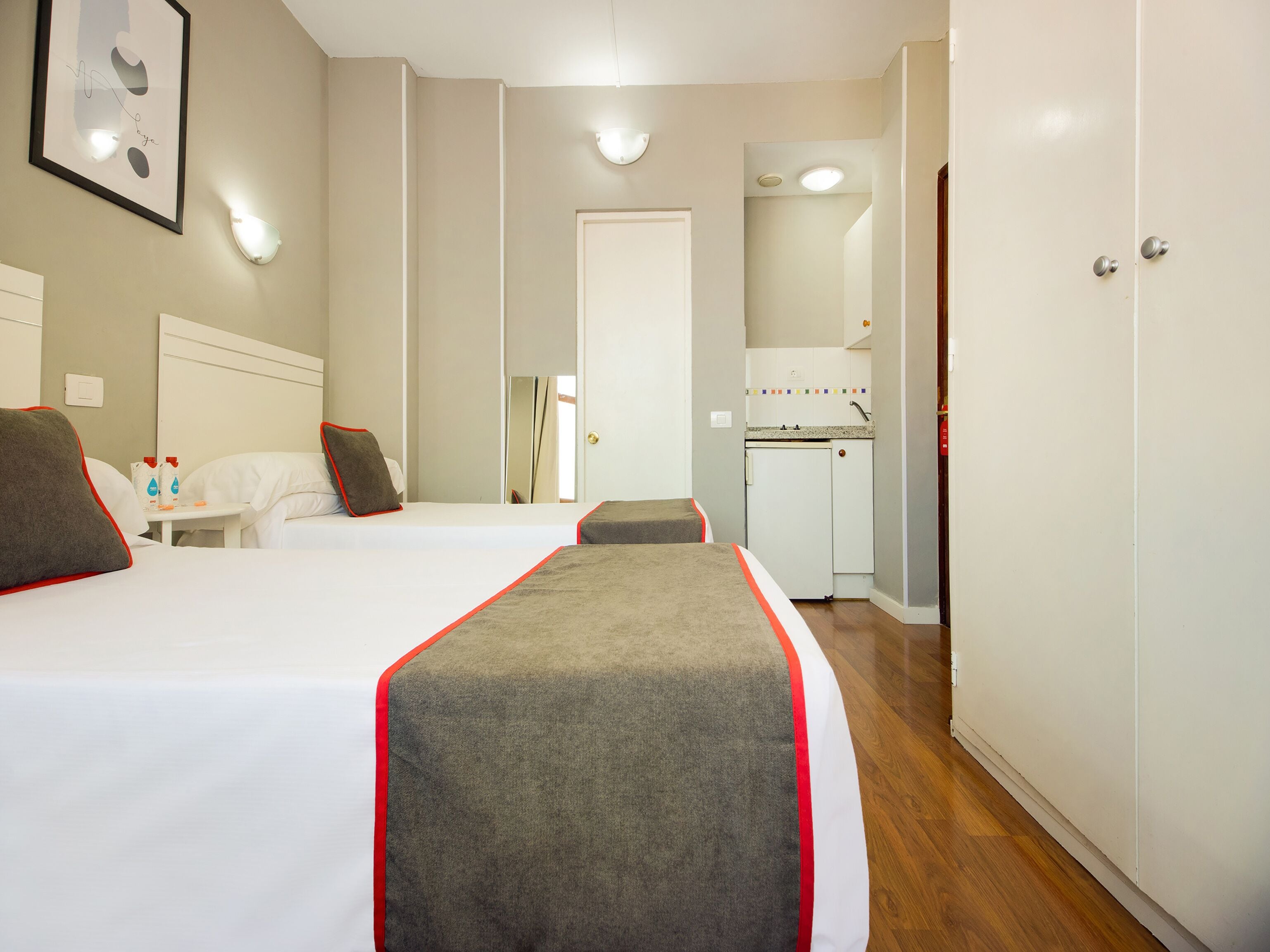 A hotel room with two beds, a small table with toiletries, a mirror, a kitchenette, and a wardrobe.