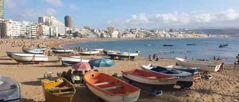 A beach with several boats and umbrellas, a sandy shore, and a cityscape in the background.