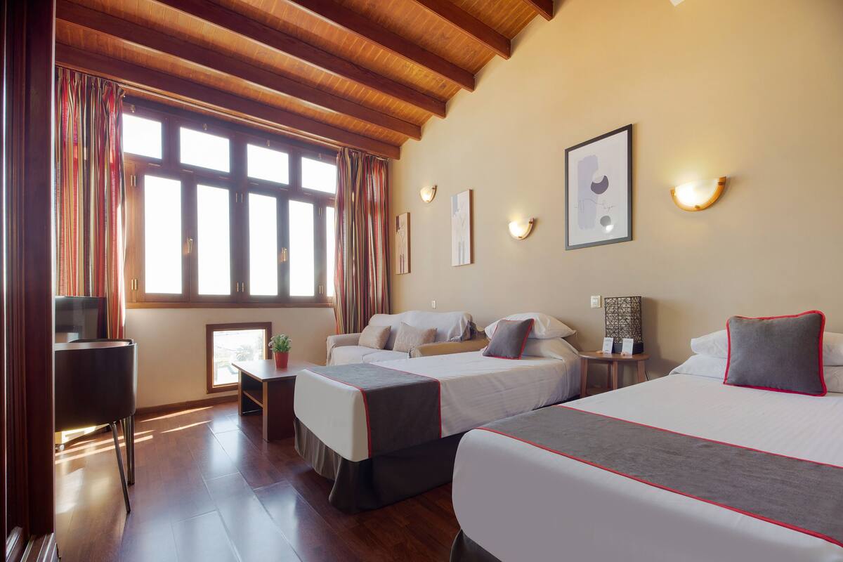 A hotel room with two beds, wooden flooring, a desk with a chair, a window with curtains, and wall-mounted lighting.