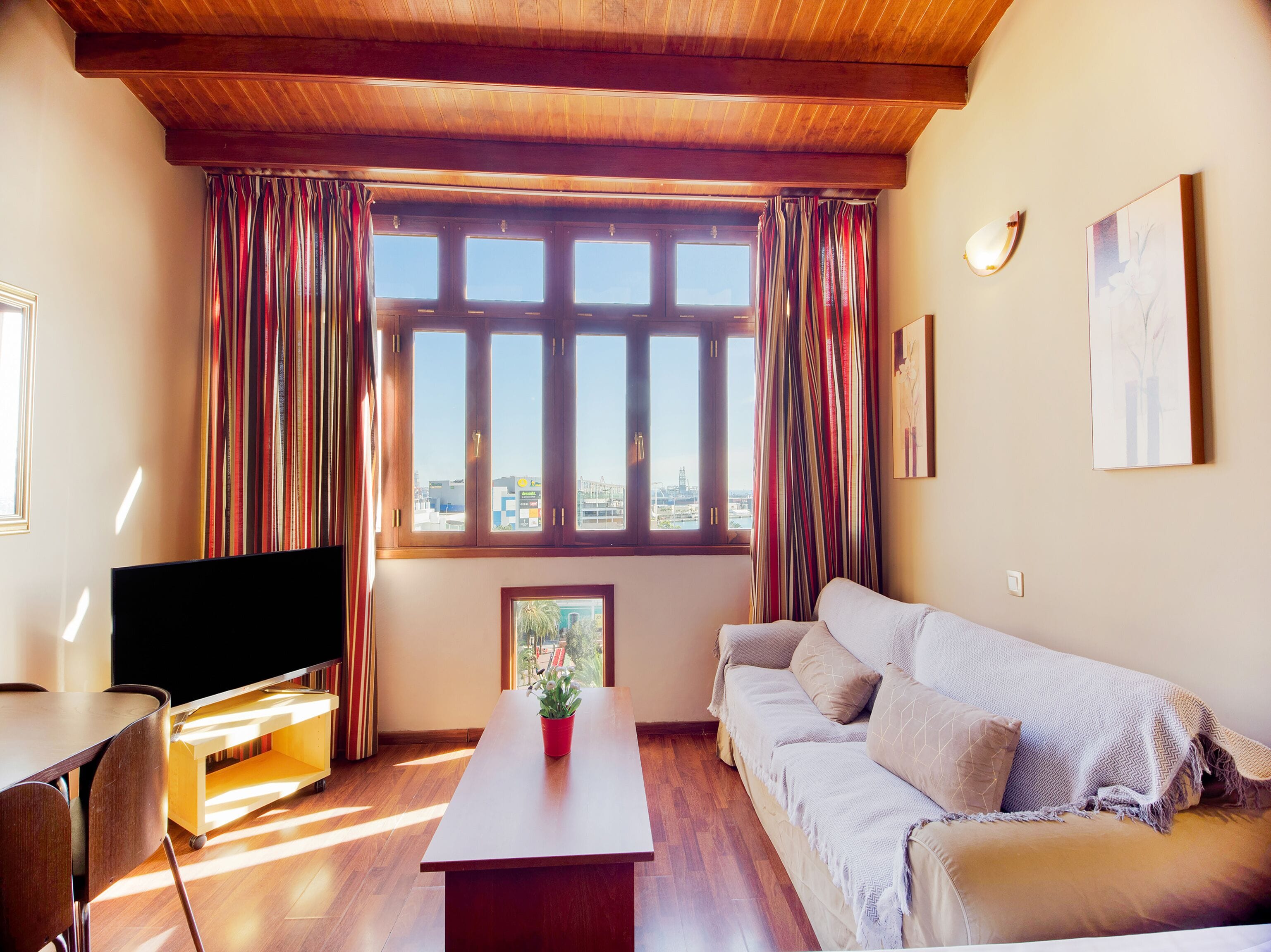 A living room with a wooden ceiling, a flat-screen TV, a sofa, a coffee table with a plant, and a window with curtains.