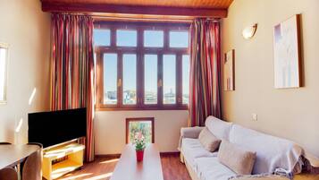 A living room with a wooden ceiling, a flat-screen TV, a sofa, a coffee table with a plant, and a window with curtains.