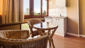 Een kamer met een eettafel en rieten stoelen, een keuken met witte kastjes en een raam met gordijnen.