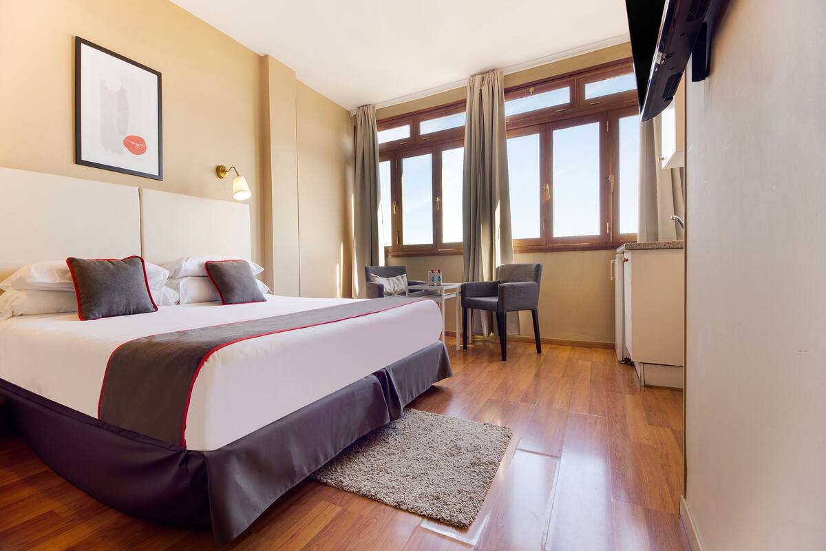 A hotel room with a large bed, a desk, and a chair. There is a window with curtains, a framed picture on the wall, and a radiator.